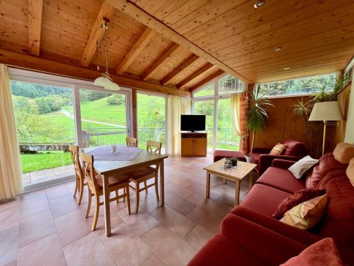 Heller Raum mit Holzdecke, großen Fenstern, einem Tisch und roter Sofagarnitur.