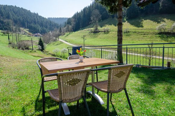 Tisch mit Stühlen auf einer Wiese, umgeben von grünen Hügeln und Bäumen.