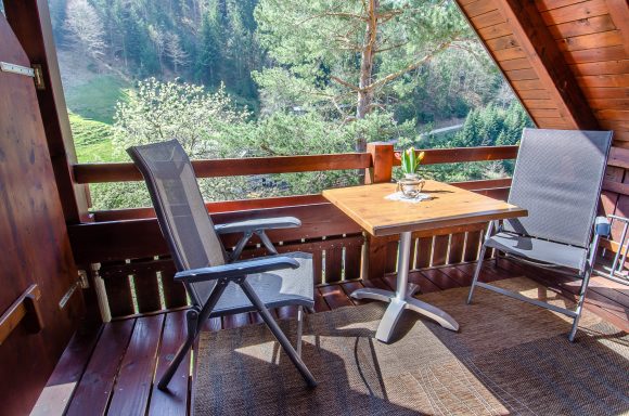 Balkon mit Tisch, zwei Stühlen und Blumen, umgeben von Bäumen und Grün.