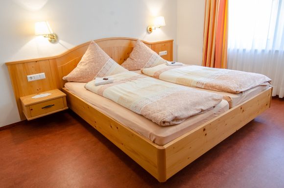 Holz Doppelbett mit zwei Matratzen und Kissen, daneben Nachttisch. Helle Vorhänge.